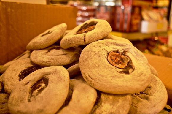 A close-up of several round, flat, grayish snacks, each with a dark, central filling, piled on top of one another inside a cardboard box. The background shows blurred shelves possibly holding other goods.