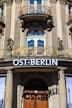 An ornate building fa&ccedil;ade features a curved balcony with intricate, gold-colored wrought iron railings. The architecture includes classical columns and decorative elements. A sign with the text 'OST-BERLIN' is prominently displayed above a large, arched wooden door that casts a shadow below.