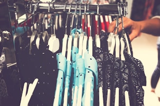A clothing rack displays a variety of shirts on hangers. The shirts feature different patterns, including polka dots and paisley prints, and appear to be in shades of blue and black. A person's hand is grasping one of the hangers, suggesting selection or browsing.