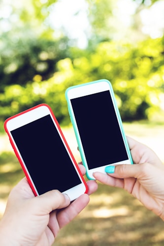 Two smartphones are held in hands against a vibrant, blurred background of green foliage. The phone on the left has a red case, while the phone on the right has a teal case. Both screens are turned off.