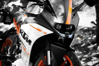 A close-up view of a sport motorcycle featuring a sleek design with prominent orange and black detailing. The angle highlights the front headlamps and part of the handlebars, with the brand logo clearly visible on the side. The background is blurred, drawing focus to the motorcycle.