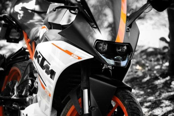 A close-up view of a sport motorcycle featuring a sleek design with prominent orange and black detailing. The angle highlights the front headlamps and part of the handlebars, with the brand logo clearly visible on the side. The background is blurred, drawing focus to the motorcycle.