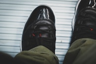 A pair of black athletic shoes with glossy sections, red detailing, and textured laces are worn with green pants. The shoes are placed on a light blue, ribbed surface, possibly a floor mat or similar material.