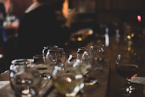 A dimly lit setting with multiple empty and partially filled snifter glasses arranged on a wooden surface. The focus is on the glasses, while the background is blurred, featuring a person sitting at a bar or table.