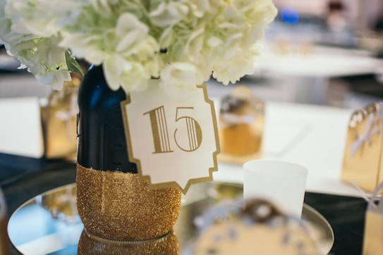 A decorative table centerpiece features a black vase with gold glitter at the base, filled with white flowers. Attached to the vase is a label with the number 15. The setup is on a reflective surface, and surrounding the centerpiece are other small decorative elements, possibly gift boxes, with a blurred background.