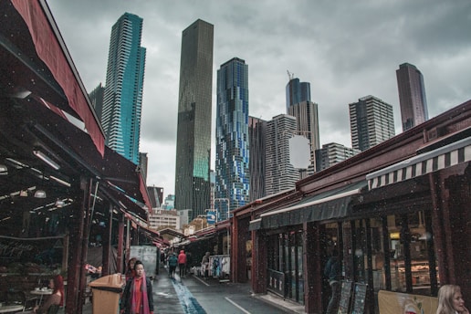 A bustling cityscape featuring a series of tall, modern skyscrapers contrasted with a lively street market below. The market stalls are lined with various goods, sheltered under red and white awnings. Overcast skies add a muted tone to the otherwise bustling scene, with people walking along the market streets.