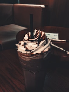 A tall glass of creamy chocolate shake next to an orange-accented napkin on a dark table.