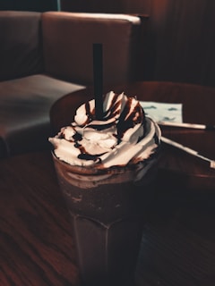A tall glass filled with a dark chocolate milkshake topped with whipped cream and chocolate syrup. The background includes a cozy setting with a couch and wooden table, giving a warm and inviting atmosphere.
