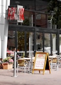 The image features an outdoor seating area of a restaurant with glass windows and a sign displaying the name 'ASK' in bold red letters. There is a sandwich board outside advertising menu items such as pizzas, pastas, and salads with their respective prices. Wicker chairs and tables are set up in the seating area, with potted flowers nearby, creating a casual dining ambiance.