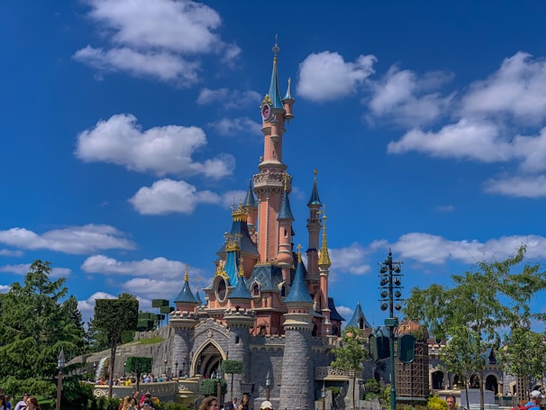 A fairy tale castle with tall spires and turrets is set against a bright blue sky with scattered fluffy clouds. The castle exhibits intricate architectural details with pastel pink and blue colors accentuated by golden trims. Lush greenery surrounds the area, and a few people are visible nearby, indicating a lively atmosphere.