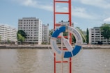 A bright blue and white lifebuoy is mounted on a red ladder by a riverside, with several modern apartment buildings in the background under a partly cloudy sky. The scene is calm, capturing an urban waterside landscape.
