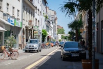 A lively street scene featuring parked and moving cars, bicycles, and pedestrians. The street is lined with shops and cafes, with signs displayed above. Palm trees and greenery add a touch of nature, while the buildings showcase classic European architecture.