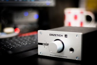 A silver-colored headphone amplifier with the brand name 'Erzetich' is prominently placed in the foreground. The device features a large control knob and several small indicators. In the background, there's a blurred keyboard and a white mug with red patterns, adding a hint of color contrast.
