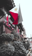 A traditional wooden building adorned with multiple Philippine flags. The structure is decorated with hanging lanterns and lush greenery along the walls, creating a rustic yet vibrant appearance. The setting appears to be an exterior view of a historical or cultural site.
