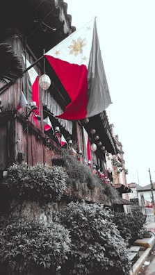 A traditional wooden building adorned with multiple Philippine flags. The structure is decorated with hanging lanterns and lush greenery along the walls, creating a rustic yet vibrant appearance. The setting appears to be an exterior view of a historical or cultural site.