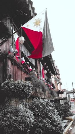 A traditional wooden building adorned with multiple Philippine flags. The structure is decorated with hanging lanterns and lush greenery along the walls, creating a rustic yet vibrant appearance. The setting appears to be an exterior view of a historical or cultural site.