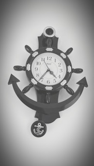 A black wall clock with a nautical theme is designed like a ship's wheel. The clock face has standard numerals and ornamental details resembling ship accoutrements. Below the clock is an anchor motif that complements the overall nautical design.