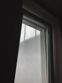 A tall, narrow window with a clear glass pane letting in natural light. The view through the window shows an adjacent plain white wall with some shadows casting across it.