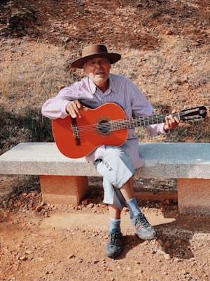 An elderly man wearing a wide-brimmed hat and a striped shirt sits on a stone bench playing an acoustic guitar. The ground is covered in rocks and dirt, with dry grass in the background, suggesting a rural setting. The lighting creates warm tones, enhancing the earthy colors.