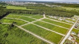 An aerial view of a green landscape featuring a grid of open grassy plots, surrounded by forested areas. Several residential houses are situated near the edges of the plots, with a larger urban area visible in the distance. The scene includes roads dividing the plots and leading into the wooded regions.