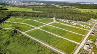 An aerial view of a green landscape featuring a grid of open grassy plots, surrounded by forested areas. Several residential houses are situated near the edges of the plots, with a larger urban area visible in the distance. The scene includes roads dividing the plots and leading into the wooded regions.