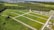 An aerial view of a green landscape featuring a grid of open grassy plots, surrounded by forested areas. Several residential houses are situated near the edges of the plots, with a larger urban area visible in the distance. The scene includes roads dividing the plots and leading into the wooded regions.