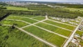 An aerial view of a green landscape featuring a grid of open grassy plots, surrounded by forested areas. Several residential houses are situated near the edges of the plots, with a larger urban area visible in the distance. The scene includes roads dividing the plots and leading into the wooded regions.