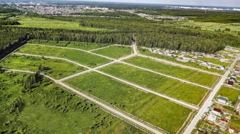 An aerial view of a green landscape featuring a grid of open grassy plots, surrounded by forested areas. Several residential houses are situated near the edges of the plots, with a larger urban area visible in the distance. The scene includes roads dividing the plots and leading into the wooded regions.