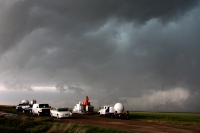 A group of vehicles parked on a rural road under a dark, ominous sky. The clouds appear heavy and stormy, suggesting severe weather conditions. Some vehicles are equipped with weather-monitoring instruments, indicating they are likely storm-chasing or studying the weather.