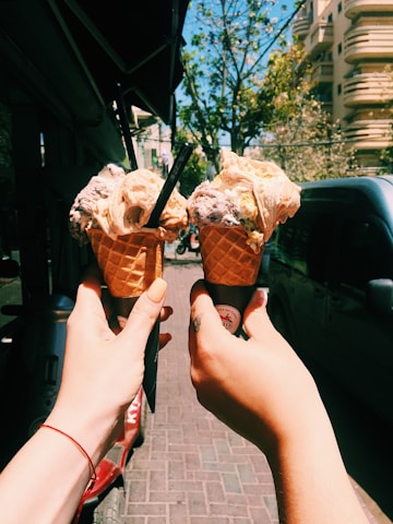 Two hands holding waffle cones filled with ice cream are raised against a sunny urban backdrop with buildings and trees lining the street. The ice cream appears to have multiple flavors and is adorned with small chocolate chips. The scene is lively and warm, suggesting a pleasant outdoor environment.
