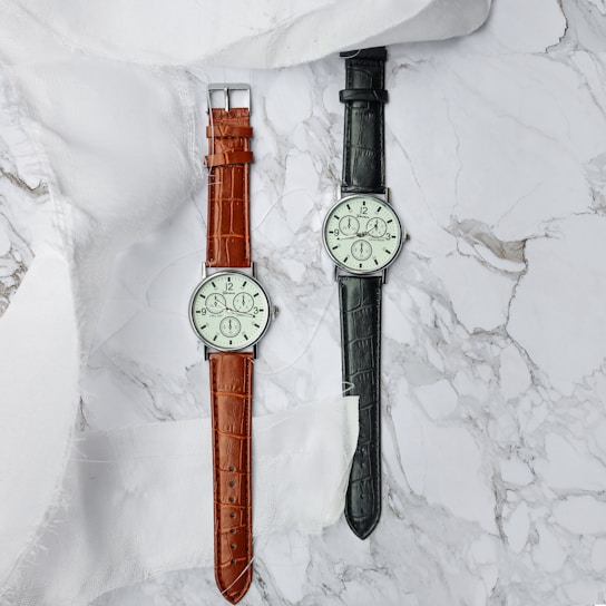 Two wristwatches are laid out on a marble surface, one with a brown leather strap and the other with a black leather strap. Both watches have classic round faces with a minimalist design, showing analog time with clear, simple numerals. A piece of sheer white fabric is draped as part of the arrangement.
