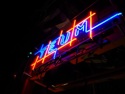 A brightly lit neon sign displaying stylized text with red and blue lights, positioned against a dark background. The neon tubes form angular, vibrant shapes with a vivid contrast to the surrounding darkness.