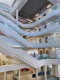A modern, multi-level interior space featuring a series of open, curved balconies overlooking a central area. The design incorporates sleek, white staircases and glass railings, providing a clean and contemporary look. Bright lighting and wide, open spaces create an airy and inviting atmosphere.