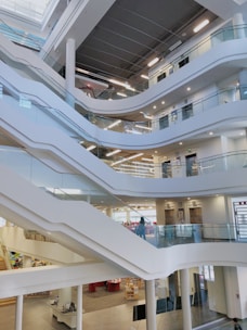 A modern, multi-level interior space featuring a series of open, curved balconies overlooking a central area. The design incorporates sleek, white staircases and glass railings, providing a clean and contemporary look. Bright lighting and wide, open spaces create an airy and inviting atmosphere.