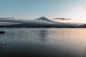 A tranquil landscape featuring a serene lake reflecting the soft hues of the sky at either sunrise or sunset. In the background, a majestic mountain, likely Mount Fuji, is partially obscured by a layer of clouds. Gentle hills surround the lake, and some buildings with lights on can be seen dotting the shore.