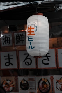 A Japanese paper lantern hangs outside a shop, displaying Japanese characters along with an illustration of a beer mug. Below the lantern, menu cards feature images of food, consisting mainly of seafood dishes decorated elegantly.