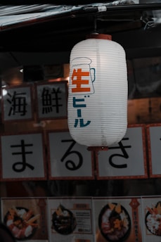 A Japanese paper lantern hangs outside a shop, displaying Japanese characters along with an illustration of a beer mug. Below the lantern, menu cards feature images of food, consisting mainly of seafood dishes decorated elegantly.
