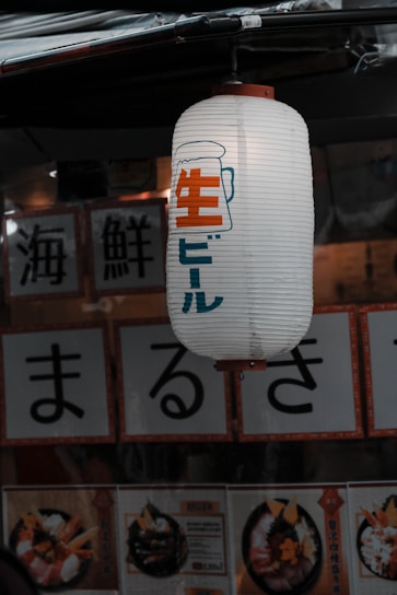 A Japanese paper lantern hangs outside a shop, displaying Japanese characters along with an illustration of a beer mug. Below the lantern, menu cards feature images of food, consisting mainly of seafood dishes decorated elegantly.