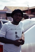 A man wearing a black hat and a light gray t-shirt with a graphic design related to 'Pinocchio the Musical' leans against a white car. The background includes blurred buildings and a slightly cloudy sky.