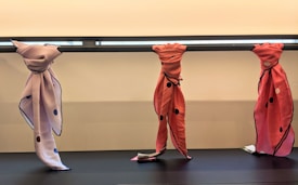 Three fabric pieces are draped and tied around a horizontal bar. The left fabric is lavender with navy blue edges and scattered circles, the middle and right fabrics are pinkish-red with black dots and edges.