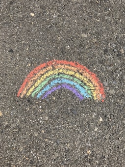 A chalk drawing of a rainbow on a rough, textured asphalt surface. The rainbow consists of vibrant colors arranged in curved stripes.