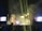 A large crowd gathers at a concert with bright stage lights and a shower of confetti falling over the audience. Large screens flank the stage, displaying colorful visuals. The atmosphere is lively, with people enjoying the event and some raising their hands in excitement.