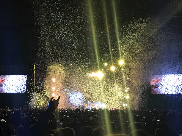 A large crowd gathers at a concert with bright stage lights and a shower of confetti falling over the audience. Large screens flank the stage, displaying colorful visuals. The atmosphere is lively, with people enjoying the event and some raising their hands in excitement.