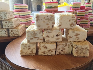 Stacks of nougat and layered jelly desserts are arranged on wooden platters. The nougat pieces are square, featuring visible nuts throughout, while the jelly desserts display colorful layered stripes.