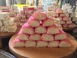 A variety of stacked sweets on wooden trays, including a prominent pyramid of pink and white coconut bars and layered nougat with nuts.