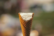 A close-up view of an ice cream cone filled with fluffy snow. The cone's texture is clearly visible, providing contrast to the smooth, snowy top. The background is blurred and features soft, muted colors, emphasizing the focus on the cone.