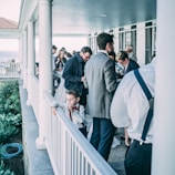 A group of people gathered on a porch, engaged in conversation and socializing. A young boy in a white shirt and red tie leans on the railing, appearing thoughtful. Adults are dressed in formal attire and are focused on the gathering.
