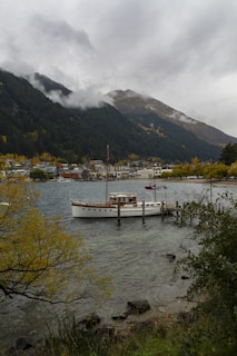 A serene lakeside scene with a wooden boat docked by a pier. The background shows a small town nestled at the base of lush, green mountains partially covered with mist. The sky is overcast with gray clouds, and the foreground features autumnal trees with yellow and green foliage along the shoreline.