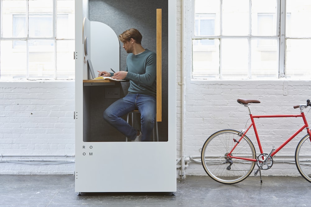 man sitting inside wooden cubicle with red bicycle parked outside