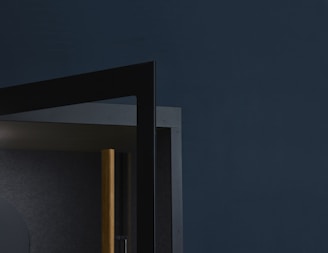 A geometric composition featuring a corner of a modern, minimalistic structure with dark, contrasting colors. The design includes a wooden element next to sleek metal frames against a deep grayish-blue background.